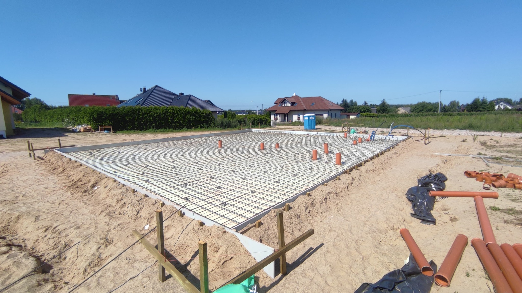 Przygotowany zbrojony fundament pod budowę domu jednorodzinnego, widoczne rury kanalizacyjne wystające pionowo, szalunki i piasek.