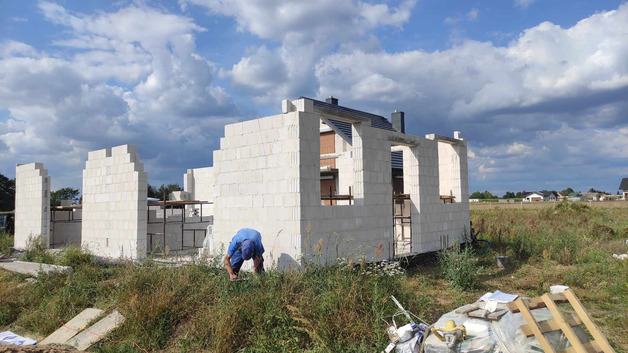 Wznoszenie ścian domu z białych bloczków, widoczny fragment dachu, pracownik w niebieskim ubraniu przy murze, trawa i nieporządek na pierwszym planie.