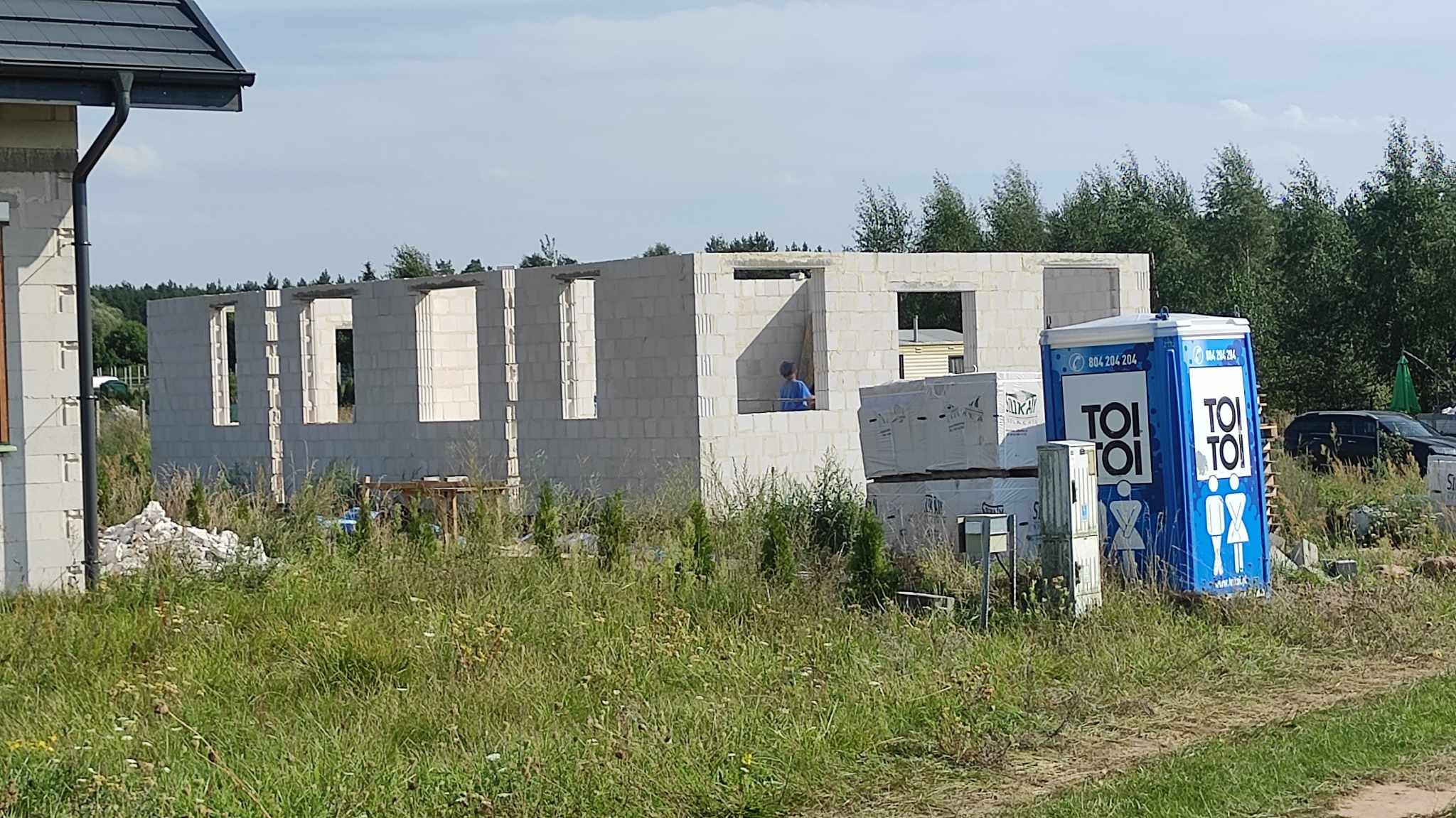 Surowy stan budynku z betonu komórkowego z widocznym pracownikiem w niebieskiej koszulce, sterta materiałów budowlanych i przenośna toaleta Toi Toi na zarośniętej działce.
