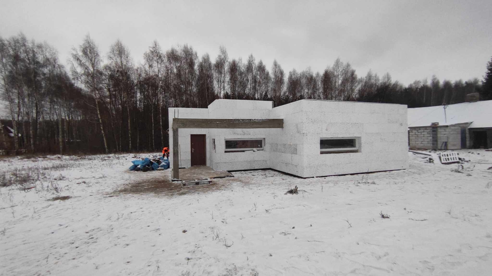 Surowy stan nowoczesnego domu z białych bloczków w zimowym krajobrazie, widoczne okna i drewniany element konstrukcyjny nad wejściem, pracownik w pomarańczowej kamizelce w tle.