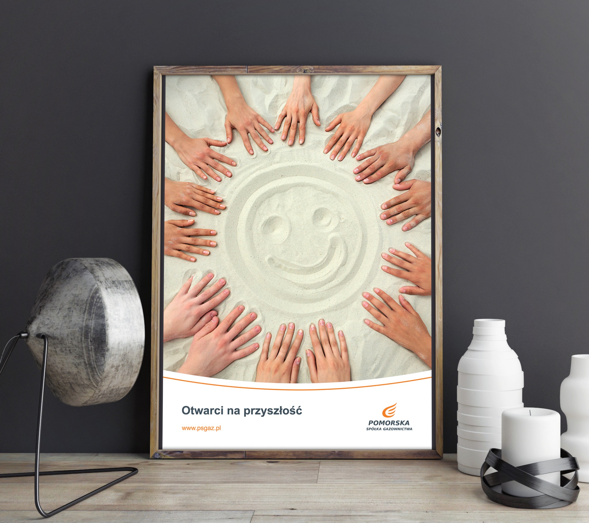 Oprawiona grafika na biurku przedstawia dłonie tworzące okrąg wokół uśmiechniętej twarzy narysowanej na piasku, z napisem 'Otwarcie na przyszłość' i logo Pomorskiej Spółki Gazownictwa.