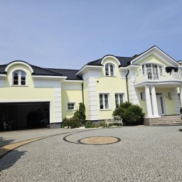 ER-BUD Eryk Stasiak - Elegancki, dwukondygnacyjny dom z żółtą elewacją, białymi detalami architektonicznymi i czarnym dachem, otoczony brukowanym podjazdem i zielenią.