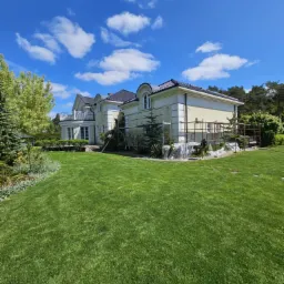 Dwukondygnacyjny dom z kremową elewacją w trakcie prac. Widoczne rusztowania i zabezpieczona folią zieleń. Słoneczny dzień, błękitne niebo z chmurami.