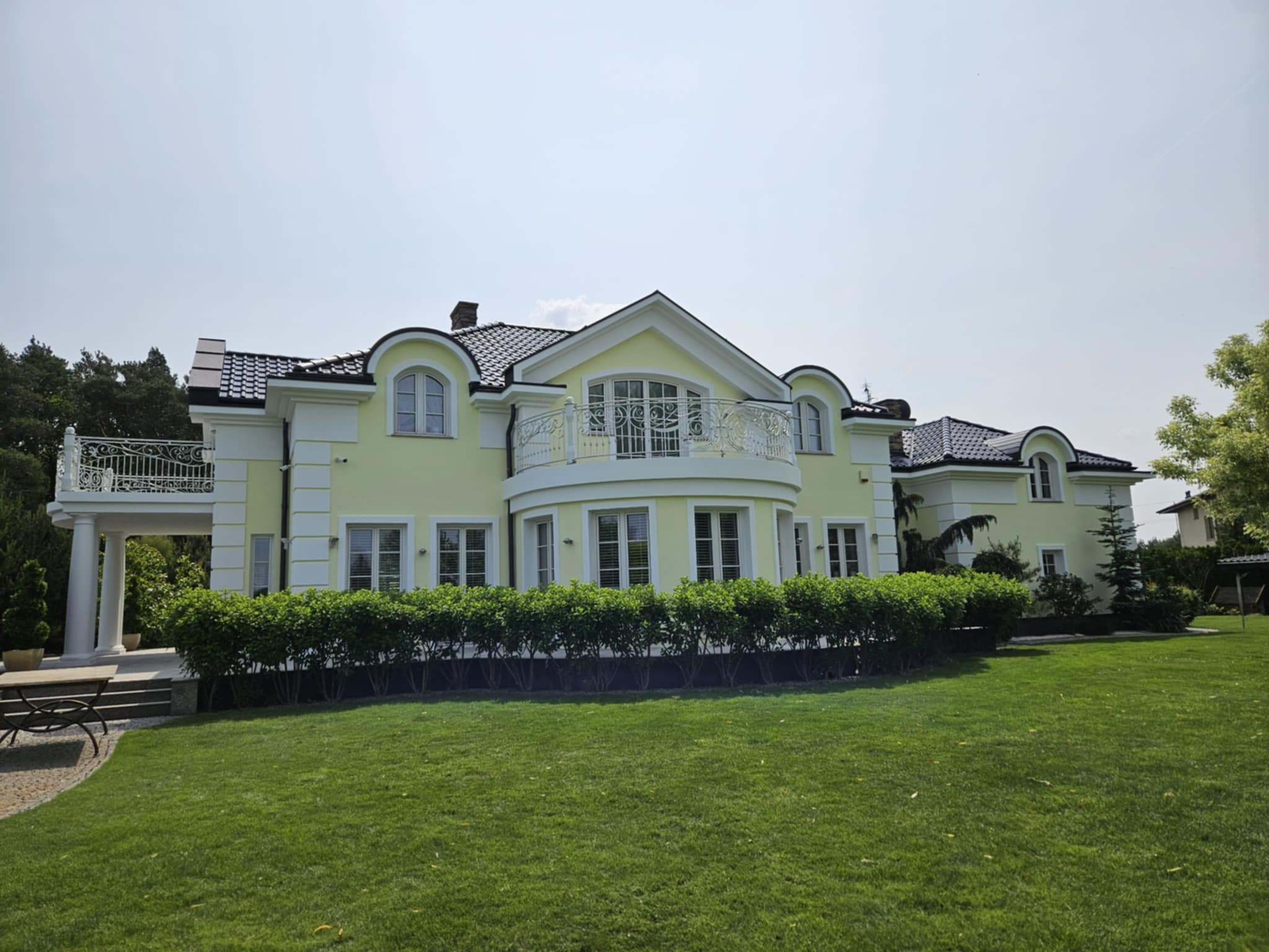 Elegancki, pastelowy dom z balkonami ozdobionymi kutą balustradą, otoczony zielonym trawnikiem i niskimi krzewami. Widok z perspektywy trawnika w słoneczny dzień.