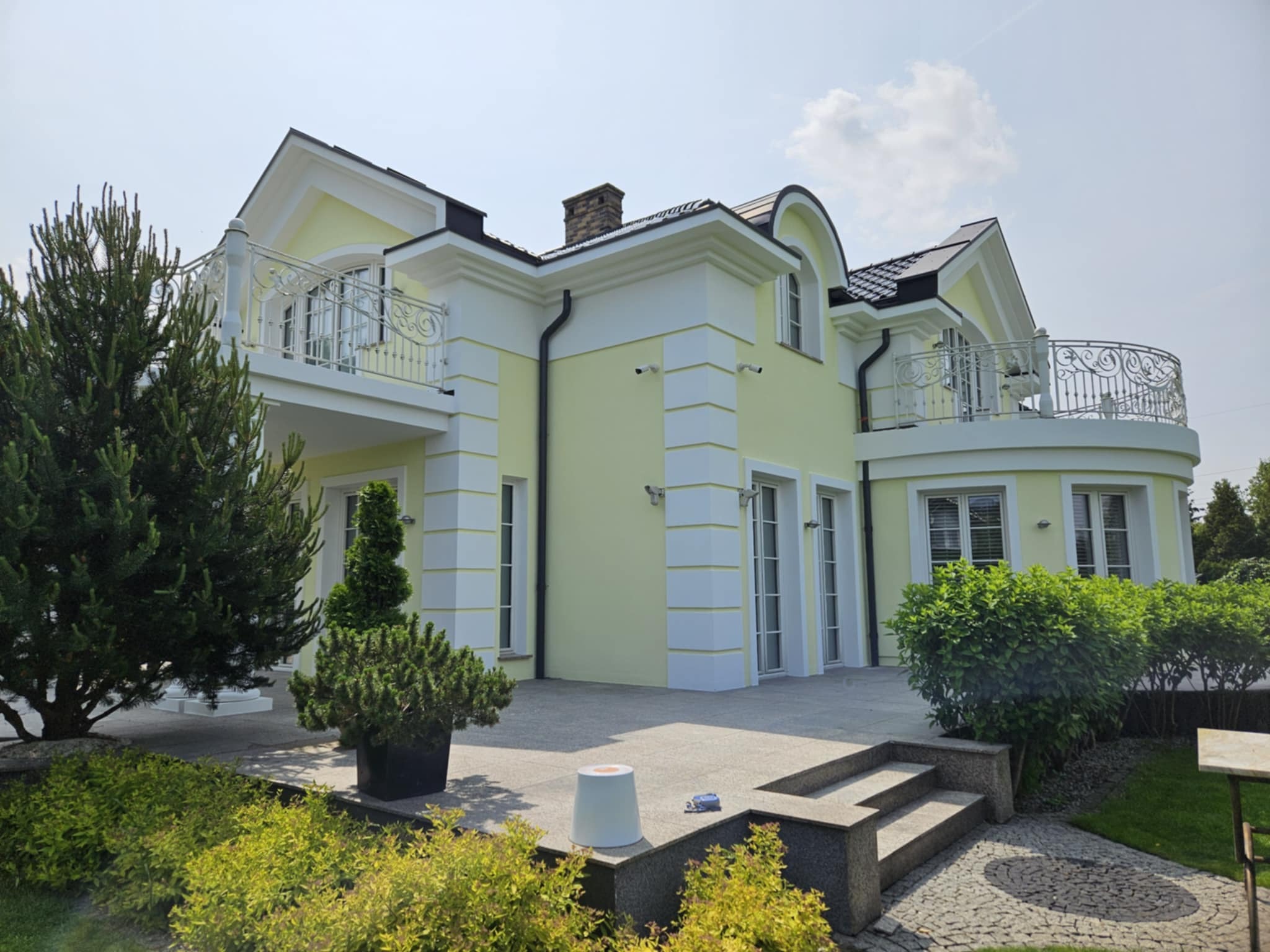 Elegancki, dwukondygnacyjny dom z pastelową elewacją i balkonami z kutą balustradą. Widoczne elementy dekoracyjne i zadbany ogród. Jasne, słoneczne oświetlenie.