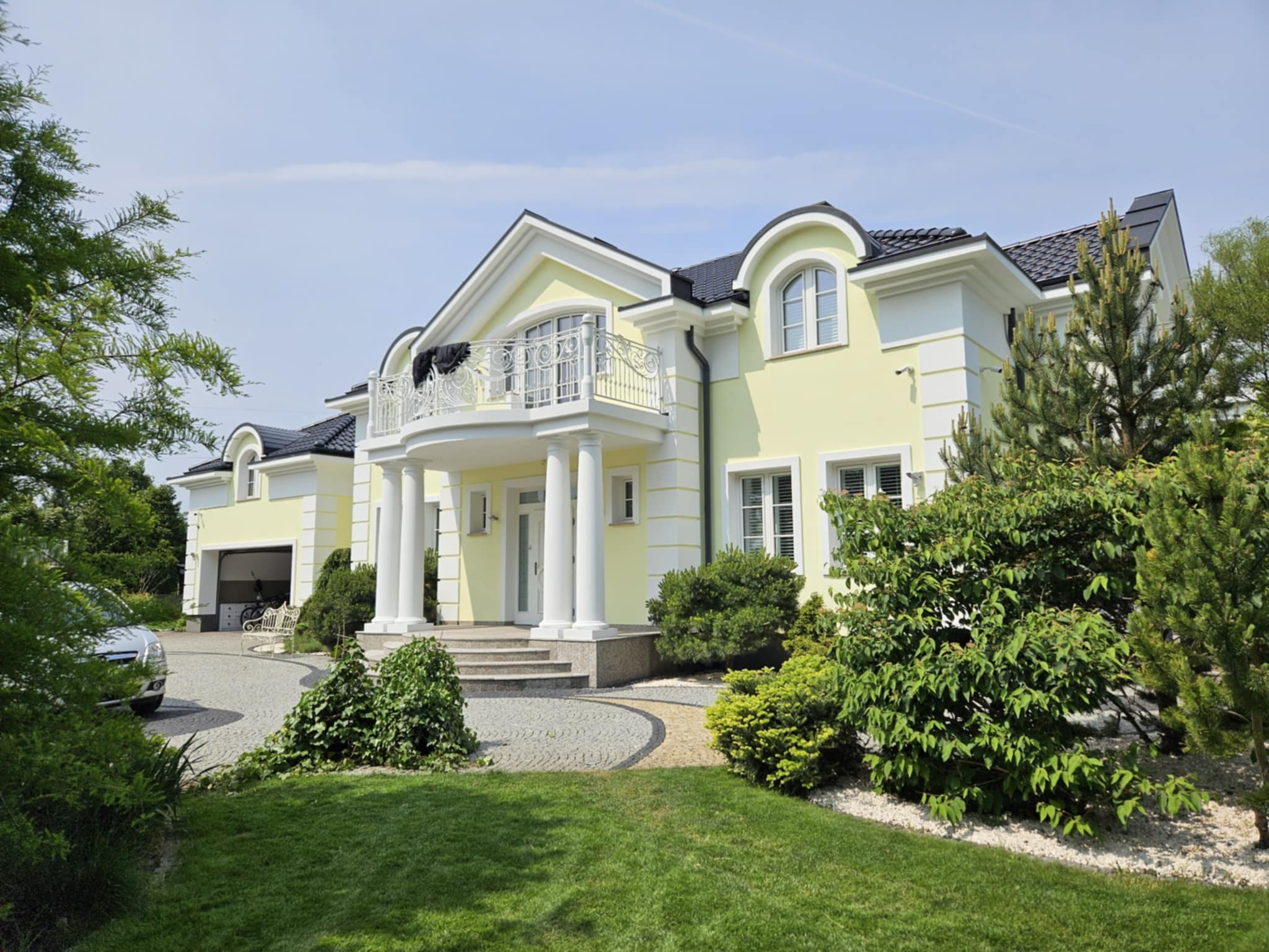 Jasnożółty dom z balkonem i kolumnami w stylu klasycystycznym, z czarnym dachem, otoczony zielenią. Widoczny podjazd wyłożony kostką.