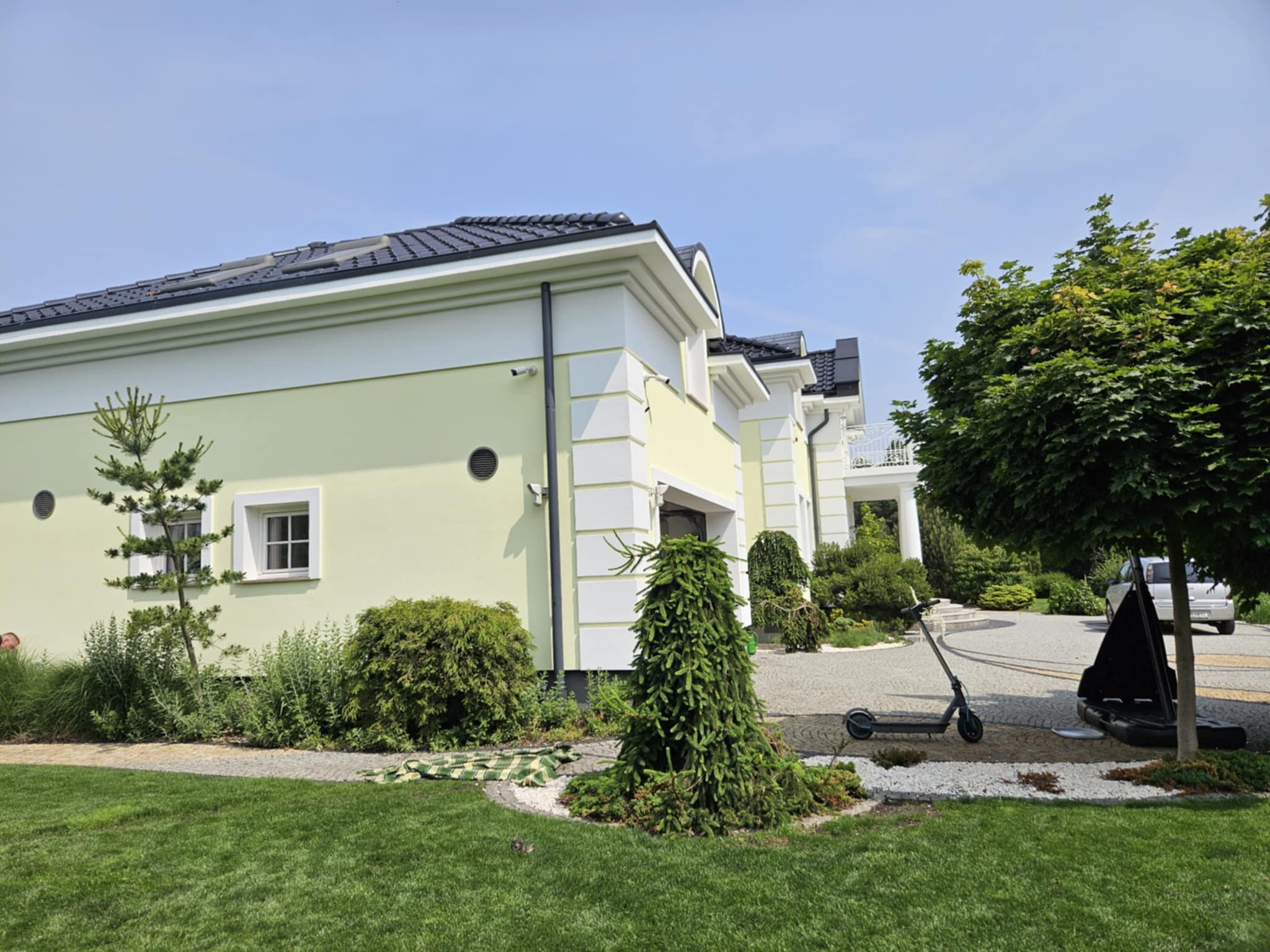 Jasnożółty dom jednorodzinny z białymi detalami architektonicznymi, czarnym dachem i zadbanym ogrodem. Widoczny fragment podjazdu i zaparkowany skuter.