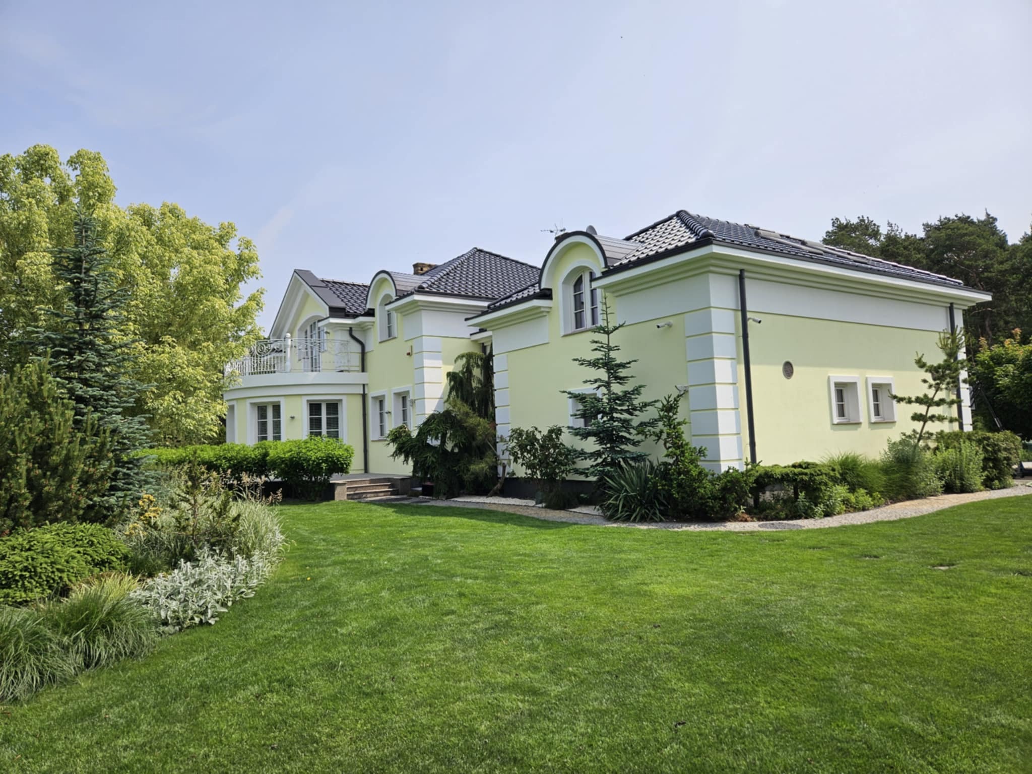 Elewacja domu jednorodzinnego w pastelowych barwach z czarną dachówką, otoczona zielenią i starannie utrzymanym trawnikiem w słoneczny dzień.