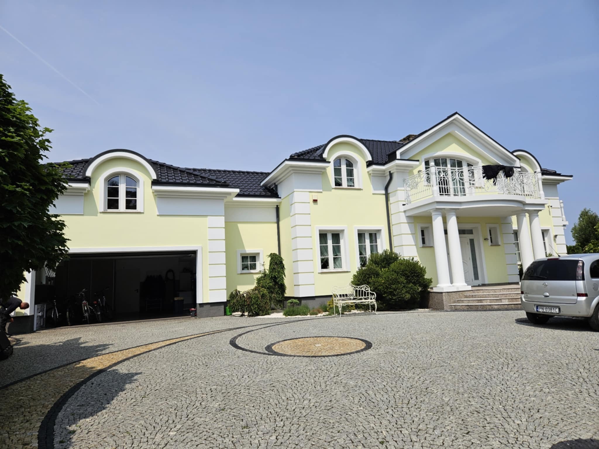 Elegancki, dwukondygnacyjny dom z żółtą elewacją, białymi detalami architektonicznymi i czarnym dachem, otoczony brukowanym podjazdem i zielenią.