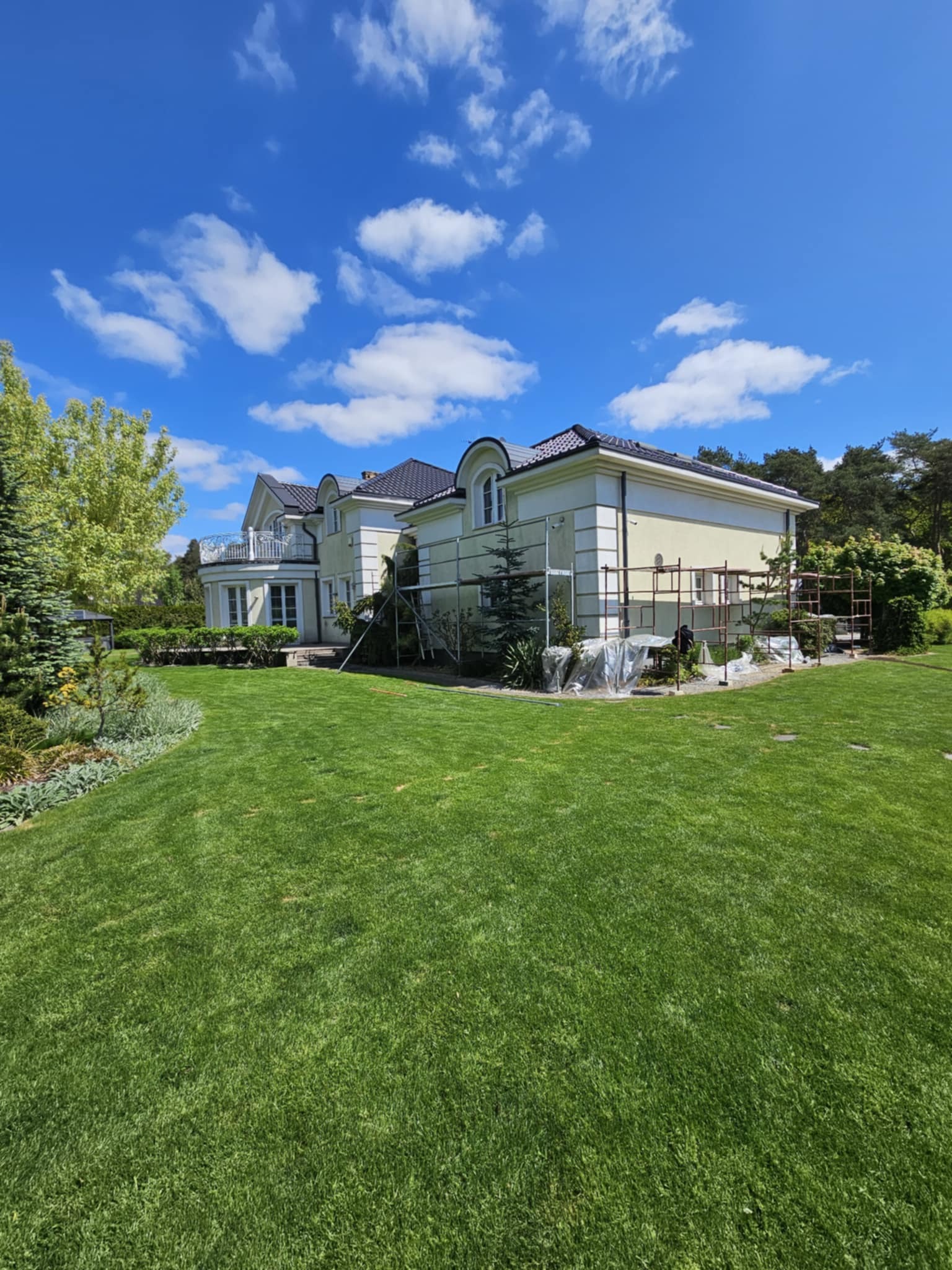 Dwukondygnacyjny dom z kremową elewacją w trakcie prac. Widoczne rusztowania i zabezpieczona folią zieleń. Słoneczny dzień, błękitne niebo z chmurami.