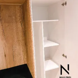 Wnętrze szafy z siedziskiem: część z półkami w kolorze białym, druga z drewnopodobną okleiną i czarnym siedziskiem. Widoczne zawiasy i logo firmy.