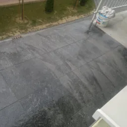 Ułożone, ciemnoszare płytki na zewnątrz, widok z góry. W tle zieleń trawy i krzewów. Wiadro z akcesoriami do czyszczenia płytek w rogu zdjęcia.