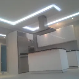 Nowoczesna kuchnia z białymi, błyszczącymi szafkami, wyspą kuchenną z czarnym blatem i oświetleniem LED w suficie, widziana z niskiego kąta.