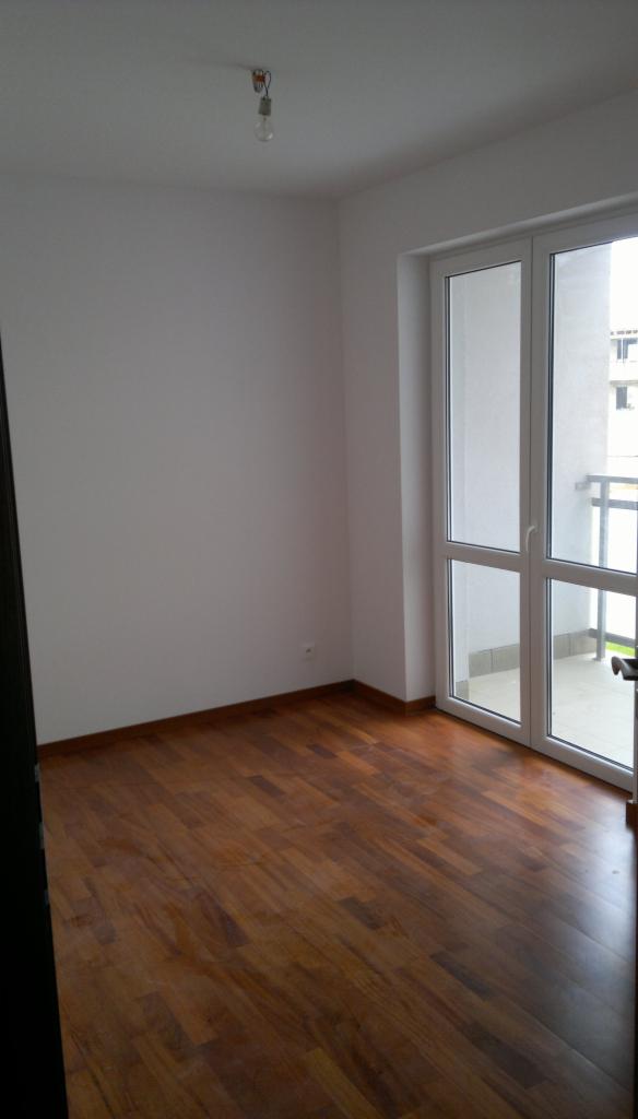 Pusty pokój po remoncie: parkiet z ciemnego drewna, białe ściany i okno balkonowe z widokiem na balkon. W suficie zwisa żarówka bez klosza.