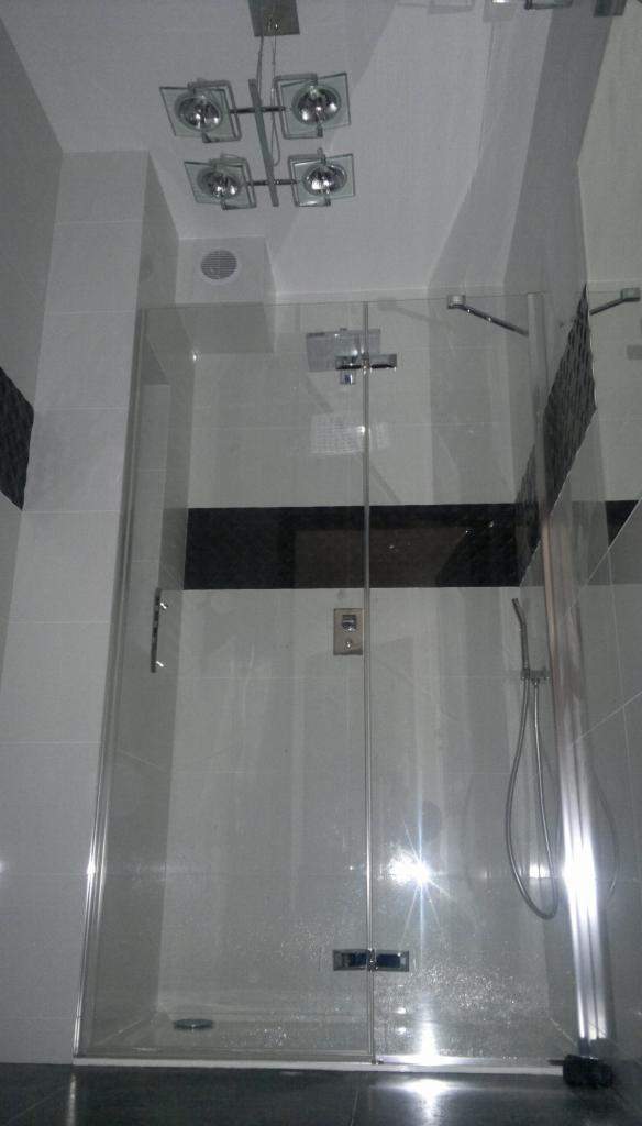 Nowoczesna kabina prysznicowa z transparentnego szkła, wykończona czarnymi akcentami, w łazience z jasnymi płytkami i designerską lampą sufitową.