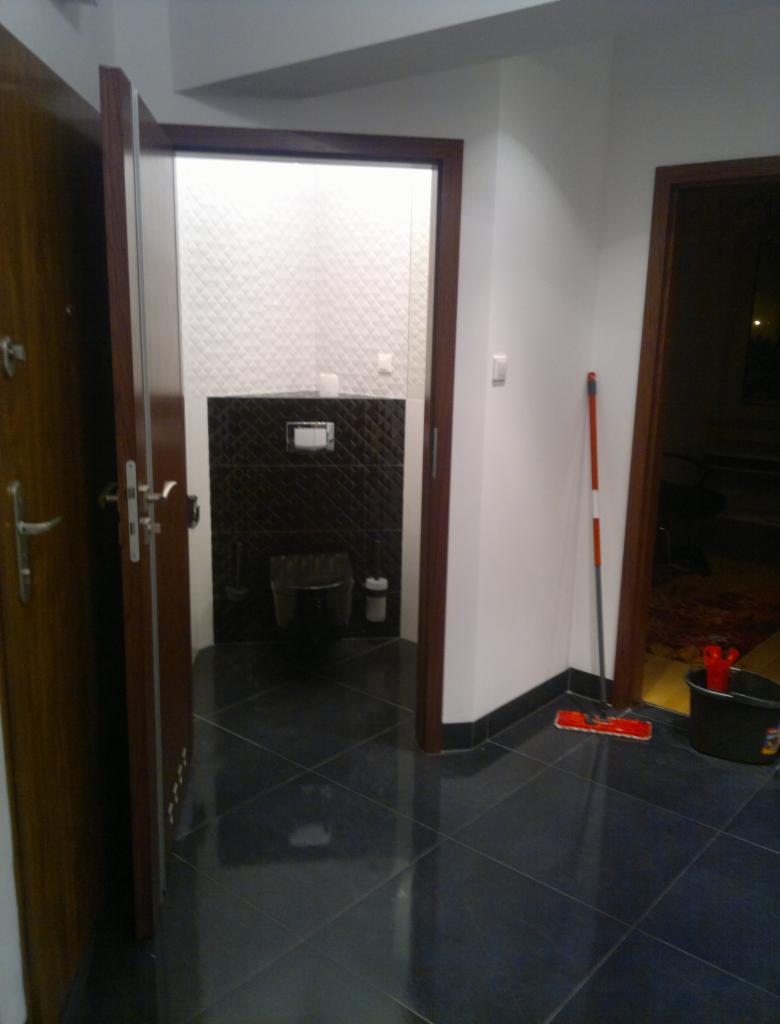 Wykończona łazienka z ciemnymi płytkami podłogowymi i częściowo wyłożoną ścianą. Widoczny mop i wiadro, sugerujące świeże sprzątanie po remoncie.