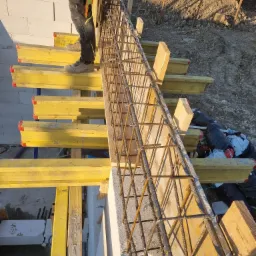 Zbrojenie szalunku wieńca na murze z betonu komórkowego, widoczne żółte stemple szalunkowe i fragment sylwetki pracownika w odzieży roboczej.