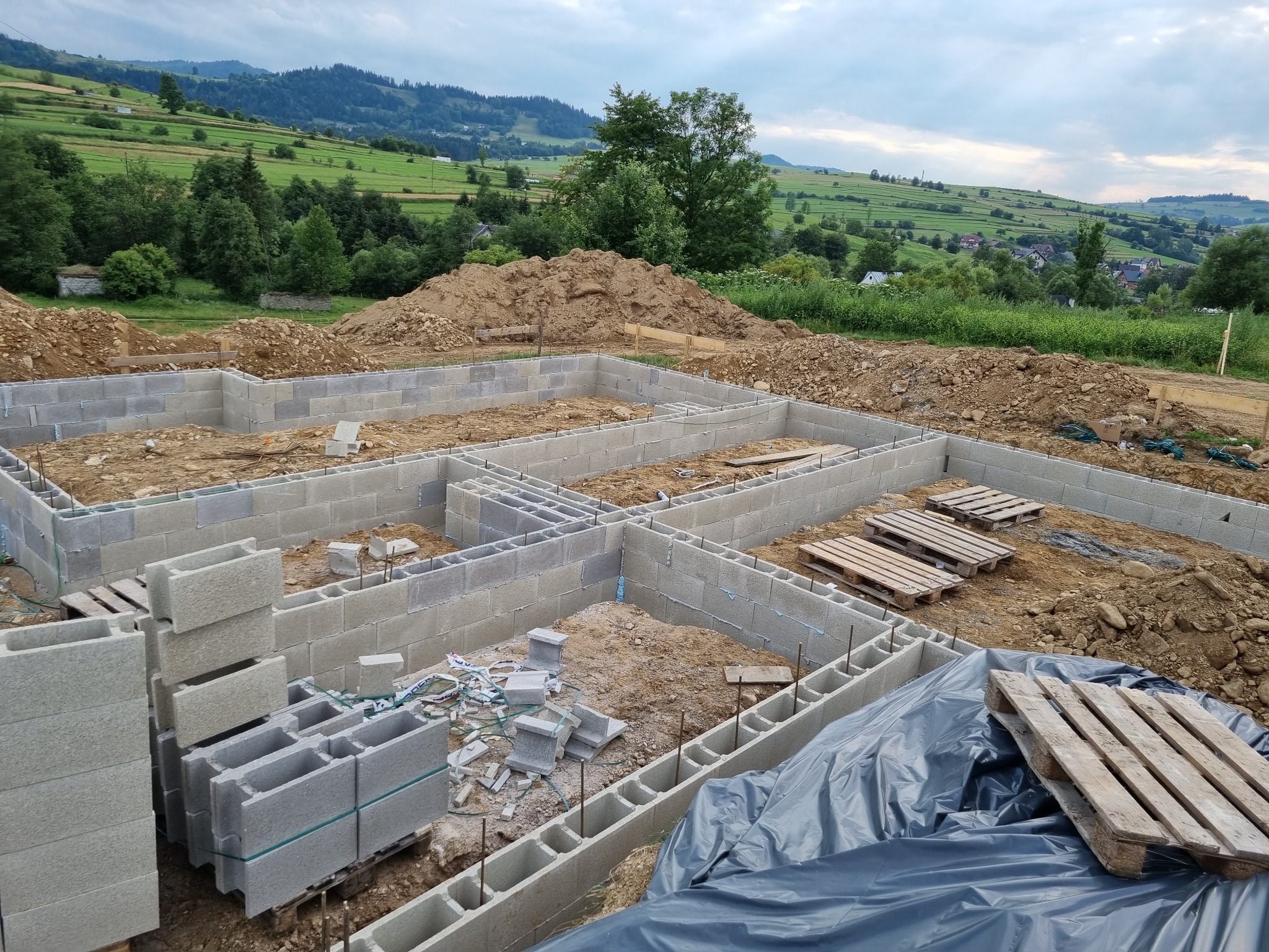 Wznoszenie fundamentów z bloczków betonowych na tle malowniczego krajobrazu z pagórkami i lasami. Część fundamentów zabezpieczona folią, widoczne palety i zapas bloczków.