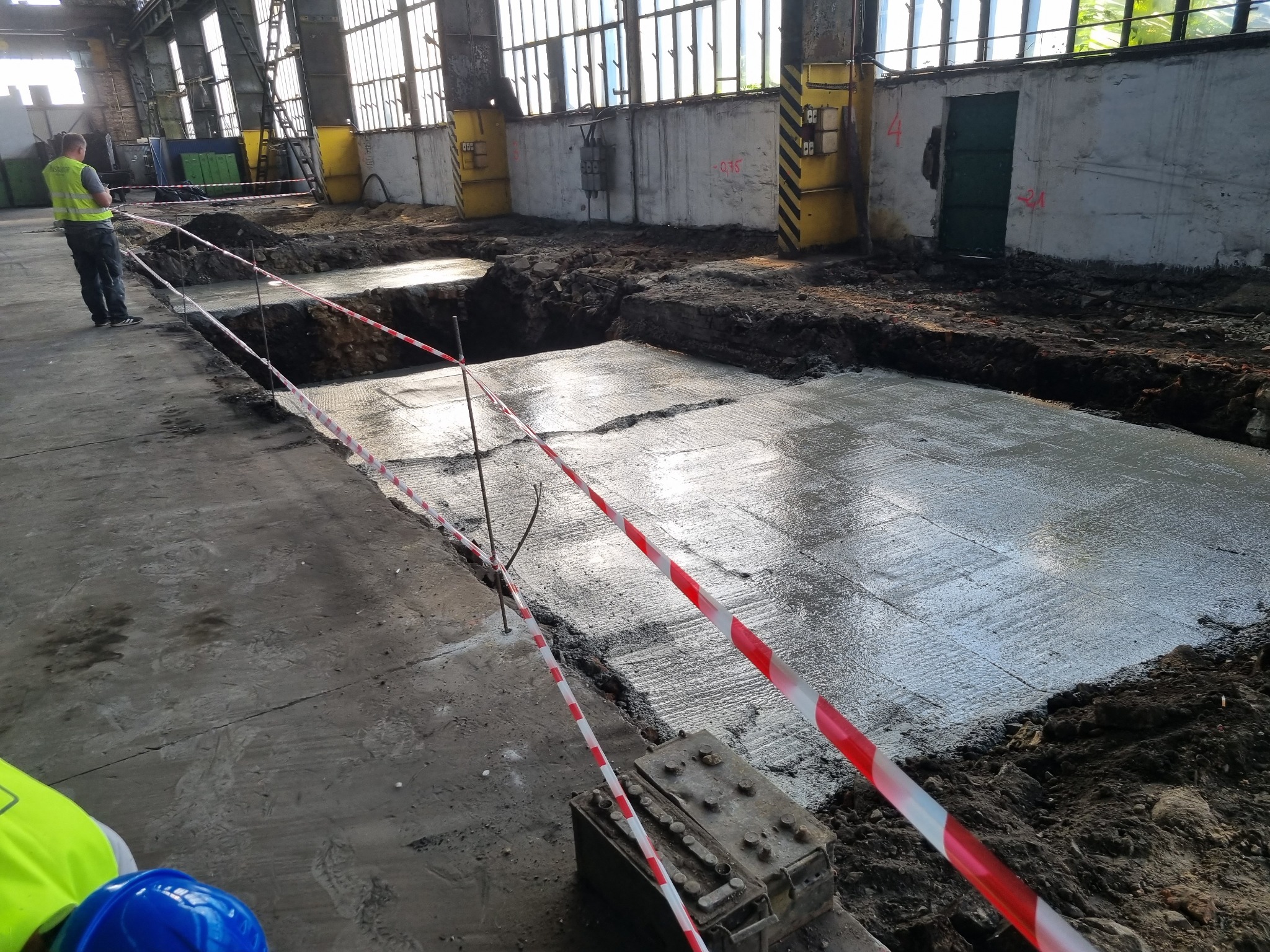 Wnętrze hali w Tychach z przygotowanym wykopem i wylanym betonem pod fundament. Widoczne taśmy ostrzegawcze i pracownik w odblaskowej kamizelce.