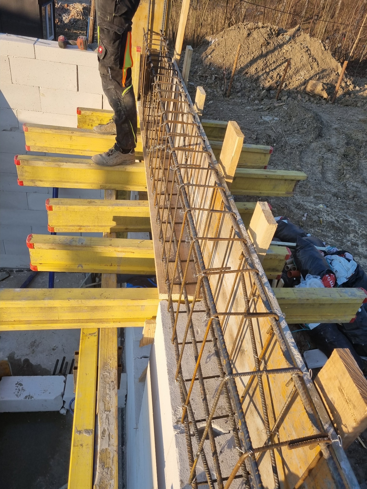 Zbrojenie szalunku wieńca na murze z betonu komórkowego, widoczne żółte stemple szalunkowe i fragment sylwetki pracownika w odzieży roboczej.