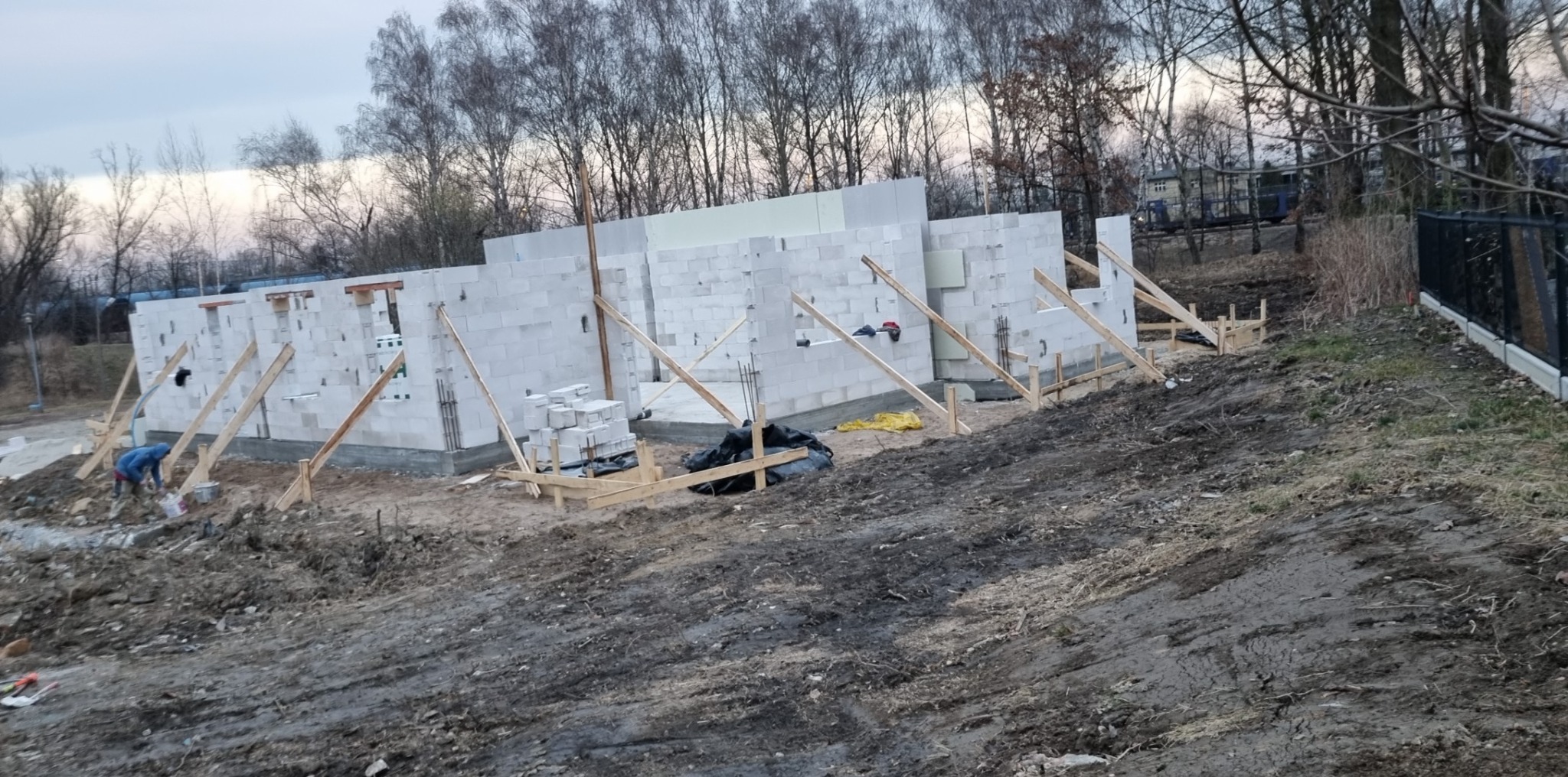 Wznoszenie ścian domu z bloczków betonowych, widoczne drewniane podpory stabilizujące, stos bloczków i pracownik w niebieskiej kurtce na placu budowy.