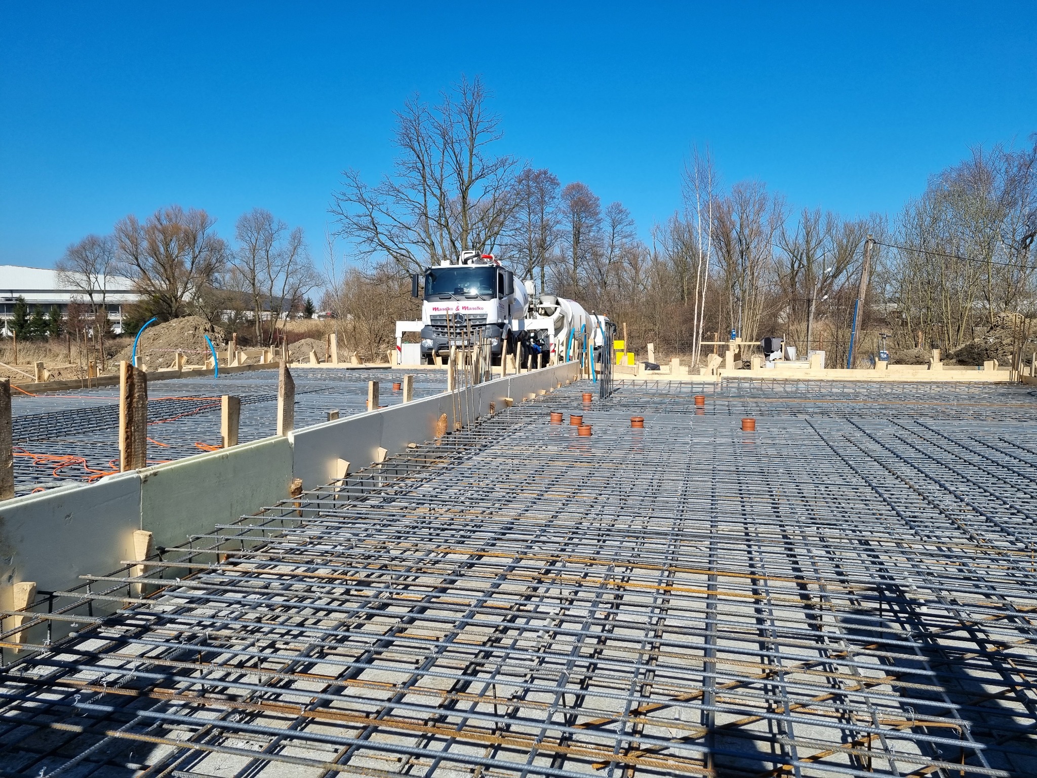 Zbrojenie fundamentu z widocznym betonowozem w tle, ujęcie w słoneczny dzień.