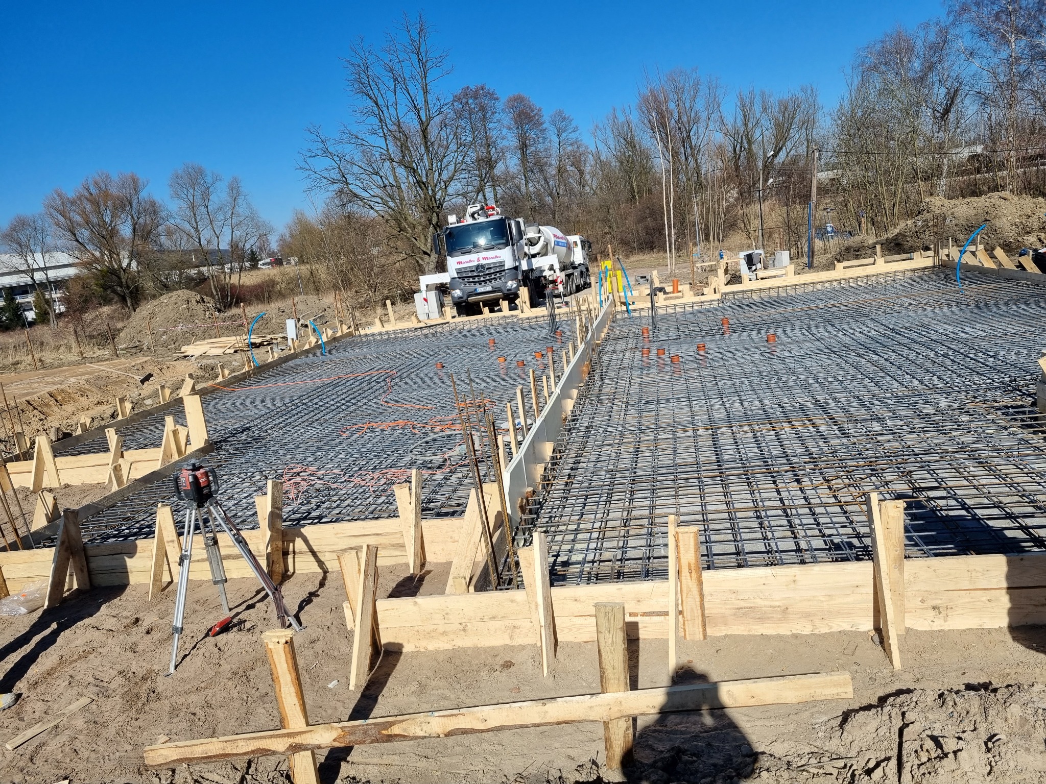 Zbrojenie płyty fundamentowej z drewnianym szalunkiem, widoczny laserowy niwelator na statywie oraz betoniarka z logo firmy w tle.