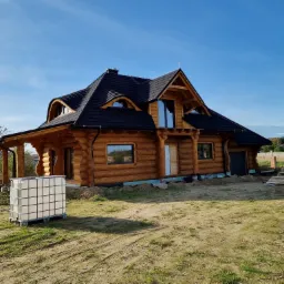 Nowoczesny dom z bali w trakcie budowy, z czarnym dachem i małymi oknami, widoczny zbiornik na wodę i materiały budowlane na placu budowy.