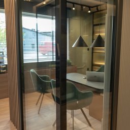 PHU DOORPLAST Piotr Jażewicz - Okno PCV z szybami zespolonymi tworzące przeszkloną ścianę biurową, widoczne biurko, krzesła i lampy wewnątrz, z zewnątrz widok na budynek mieszkalny.