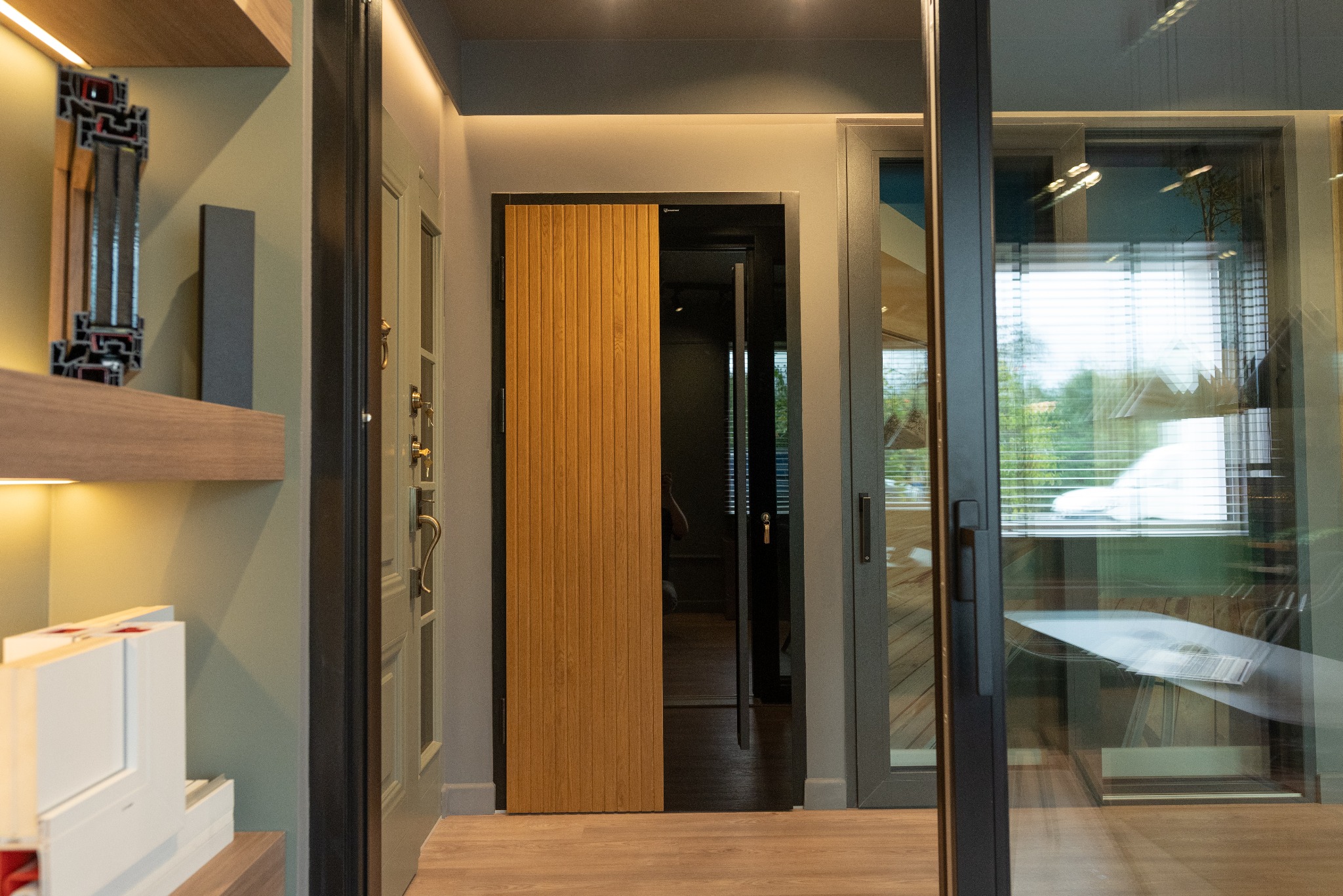 Ekspozycja wnętrza: dębowa, przesuwna płyta drzwiowa z pionowymi lamelami w czarnej ramie, częściowo zasłaniająca widok na sąsiednie pomieszczenie, obok widoczne okno przesuwne i przekrój profilu...