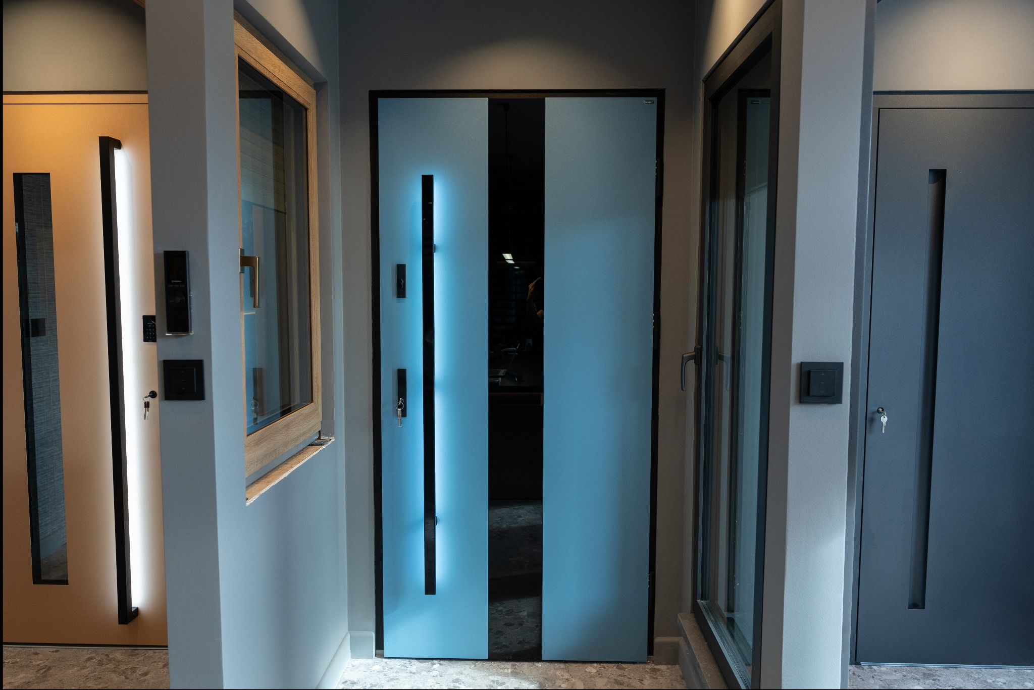 Wnętrze z trzema różnymi modelami drzwi zewnętrznych: beżowe z podświetleniem LED, błękitne z czarnym pionowym przeszkleniem i szare z prostokątnym wgłębieniem, wszystkie z zamontowanymi klamkami...