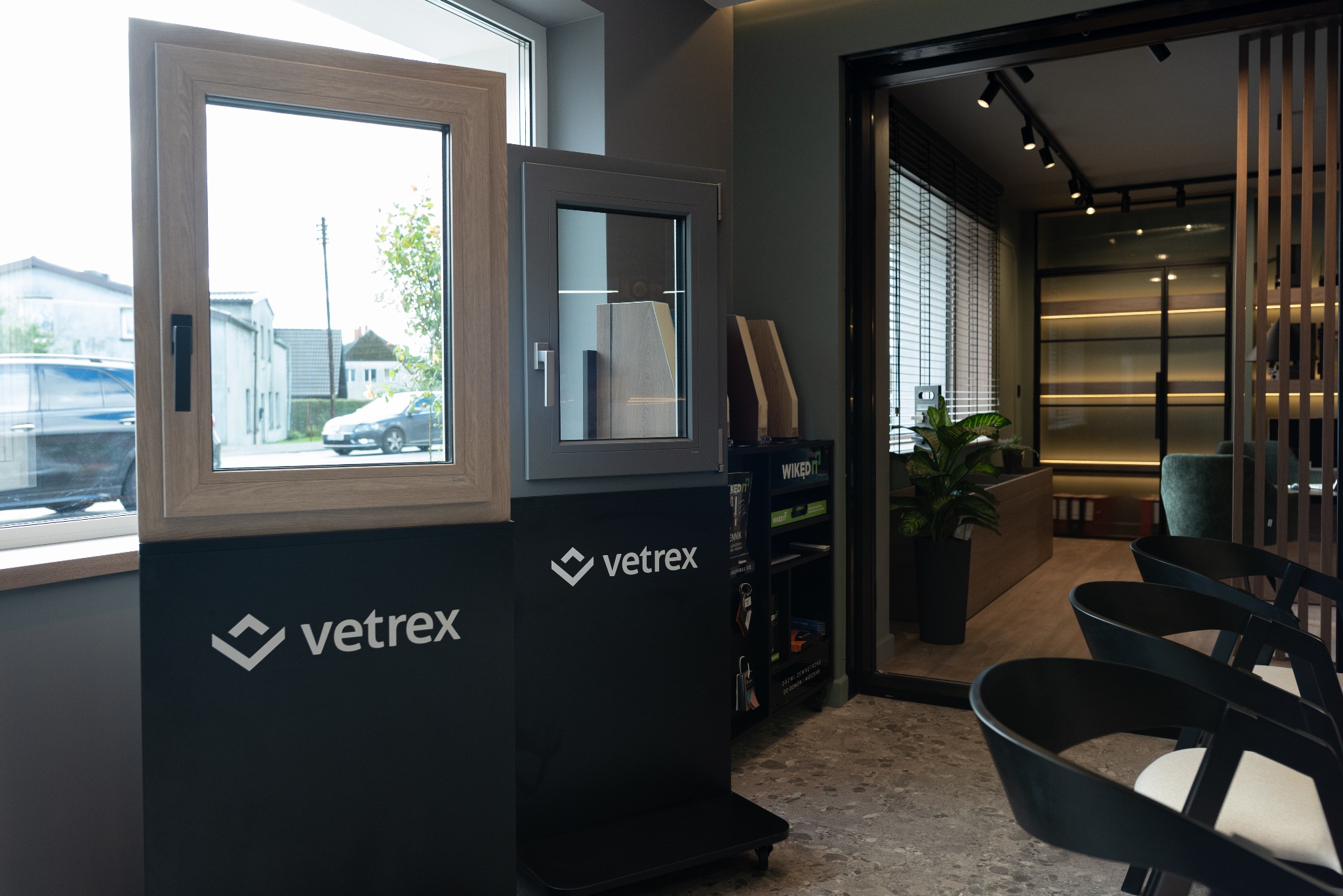 Ekspozycja dwóch modeli okien z różnymi ramami w salonie, widoczne logo producenta na czarnym panelu, w tle biuro z żaluzjami i rośliną.