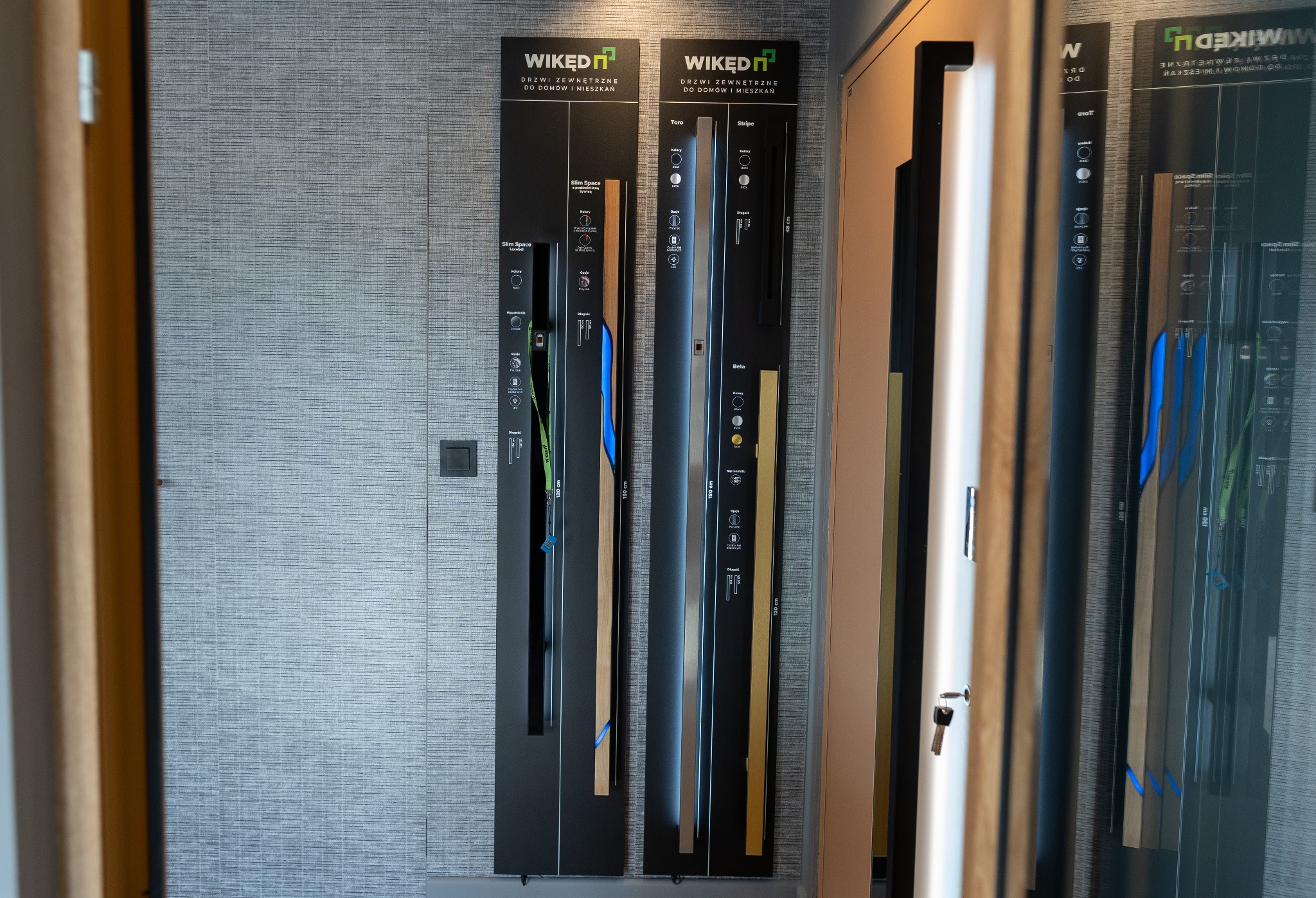 Ekspozycja modeli drzwi zewnętrznych firmy Wikęd na czarnych panelach informacyjnych z opisami, widoczne fragmenty drzwi w różnych kolorach i wykończeniach, w tle tapeta o strukturze lnu.