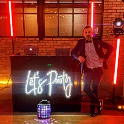 Mężczyzna w garniturze z muszką stoi oparty o podświetlony panel DJ'ski z napisem 'Let's Party', obok dekoracyjny lampion i czerwone lampy, w tle ceglana ściana.