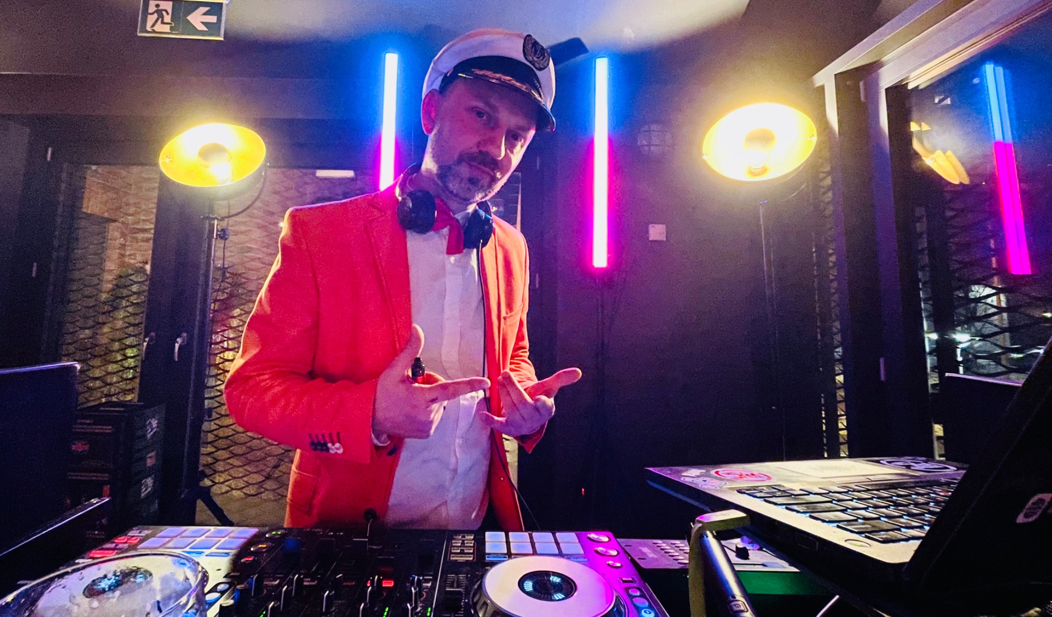 DJ w czerwonej marynarce i czapce kapitana, z słuchawkami na szyi, prezentuje sprzęt DJ-ski podświetlony neonami i lampami punktowymi.