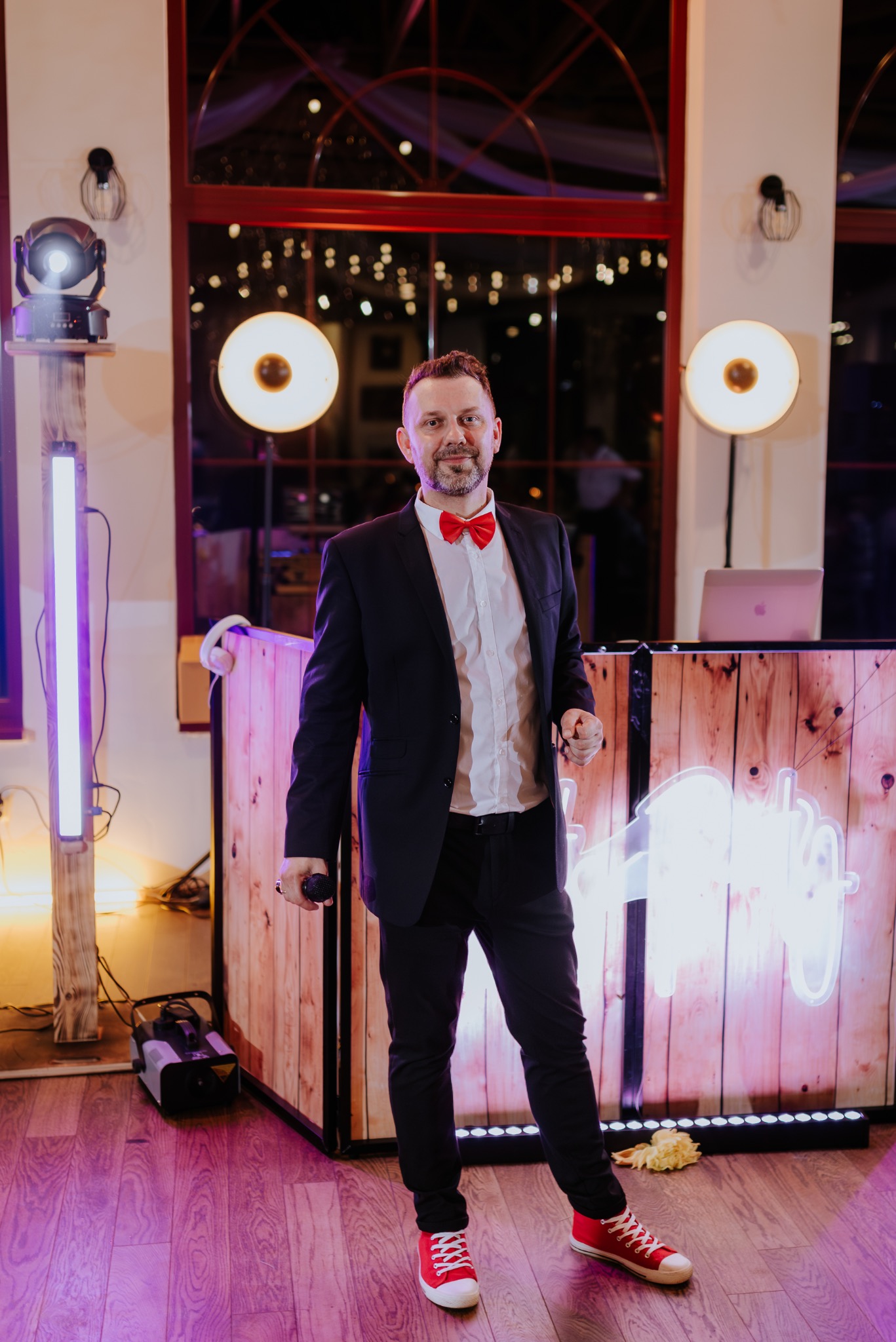 Prezenter w czarnym garniturze i czerwonej muszce, trzymający mikrofon, stojący przed drewnianą ścianką z neonowym napisem, oświetloną reflektorami i lampami LED.