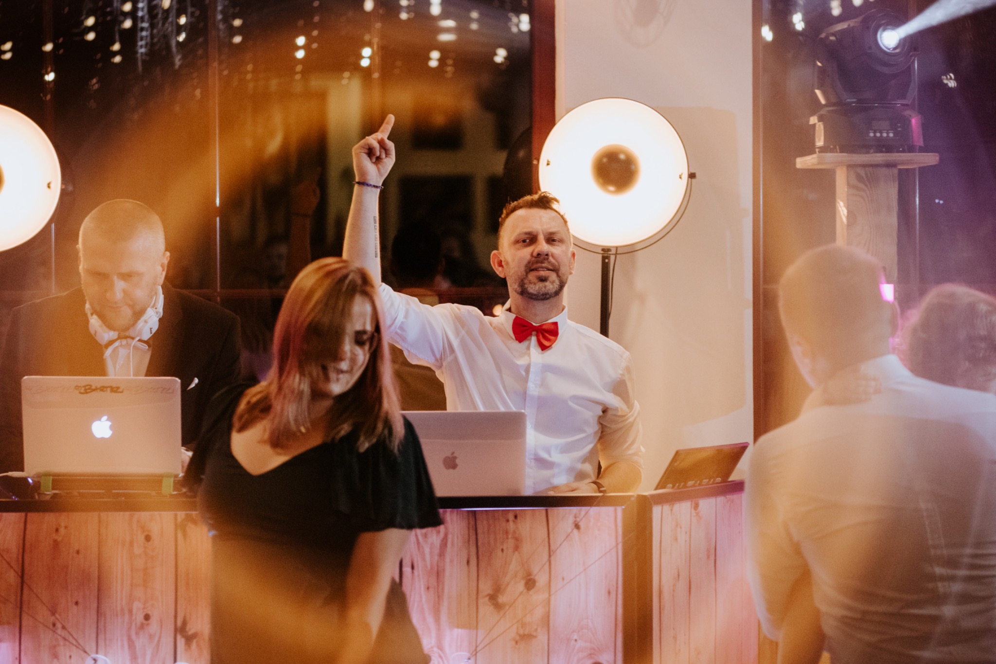 Dj w białej koszuli i czerwonej muszce, z uniesioną ręką, za drewnianym stanowiskiem z laptopem, obok drugi Dj z laptopem i słuchawkami, oświetlenie sceniczne, tańczący ludzie.