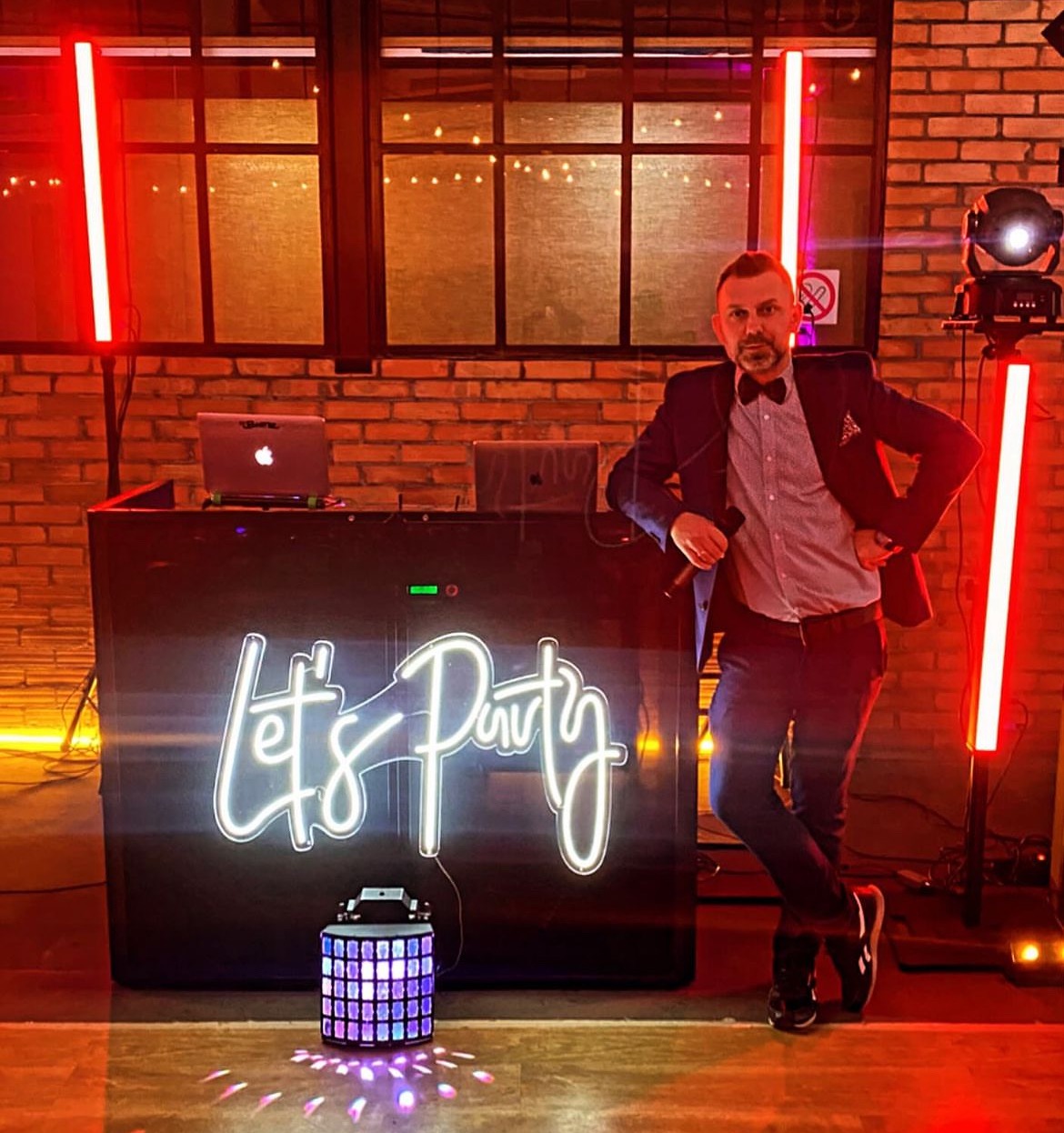 Mężczyzna w garniturze z muszką stoi oparty o podświetlony panel DJ'ski z napisem 'Let's Party', obok dekoracyjny lampion i czerwone lampy, w tle ceglana ściana.