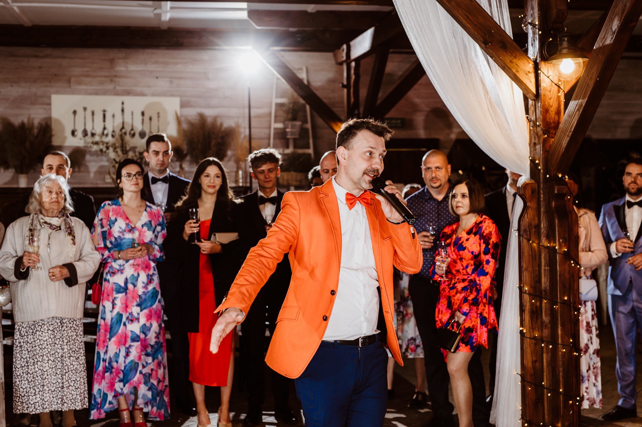Mężczyzna w pomarańczowej marynarce z mikrofonem prowadzi imprezę okolicznościową, w tle goście weselni i drewniana dekoracja z lampkami.