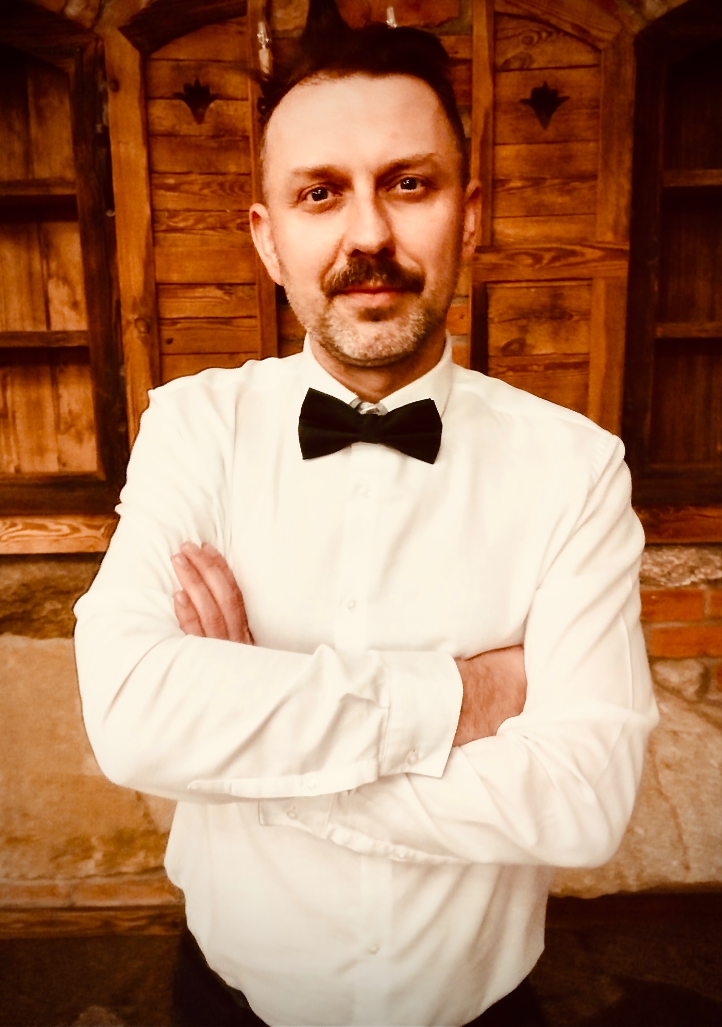 Portret mężczyzny z wąsami i muchą, stojącego na tle drewnianych drzwi i ceglanej ściany, z założonymi rękami.