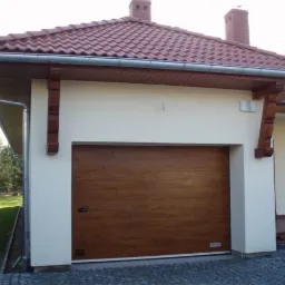 Ocieplana segmentowa brama garażowa o fakturze drewna, zamontowana w budynku z beżową elewacją i czerwoną dach&oacute;wką, widok z zewnątrz.