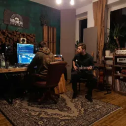 Wnętrze studia nagrań: mężczyzna pracuje przy komputerze z oprogramowaniem do edycji dźwięku, drugi gra na gitarze elektrycznej. Na ścianie dekoracyjny panel akustyczny i logo studia 'Kreacja...