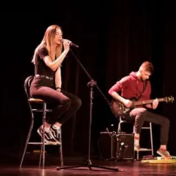 Wokalistka śpiewa do mikrofonu siedząc na wysokim krześle, obok gitarzysta w czerwonej bluzie gra na gitarze elektrycznej na scenie z czarnym tłem, na lewo od wokalistki stoi stolik z trofeami.