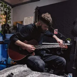 Młody mężczyzna z brodą stroi gitarę akustyczną w domowym studio, widoczne pianino, mikrofon, gitary, wytłumienie akustyczne na ścianie i rośliny.