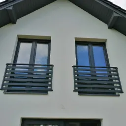 Elewacja domu z dwoma oknami zabezpieczonymi czarnymi, metalowymi balustradami balkonowymi o poziomym układzie szczebli.