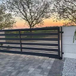 Nowoczesna, przesuwna brama wjazdowa z poziomymi panelami na tle zachodzącego słońca i dekoracyjnej roślinności.