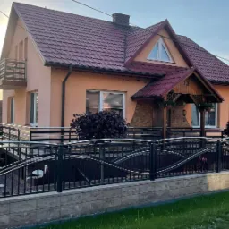 Czarna metalowa brama z ozdobnymi, falistymi elementami przed domem z pomarańczową elewacją i czerwoną dachówką, widoczne rzeźby bocianów w ogrodzie.