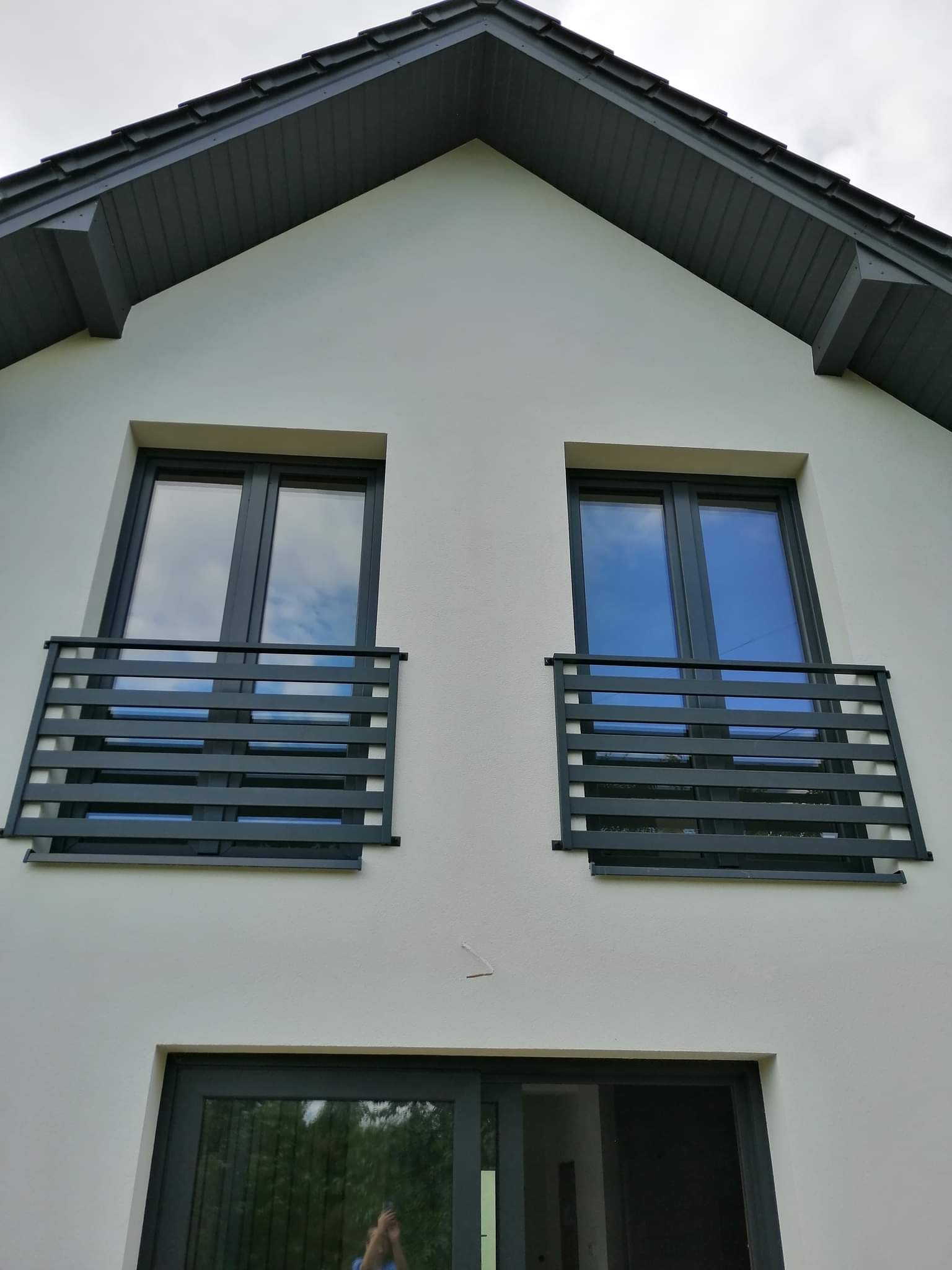 Elewacja domu z dwoma oknami zabezpieczonymi czarnymi, metalowymi balustradami balkonowymi o poziomym układzie szczebli.