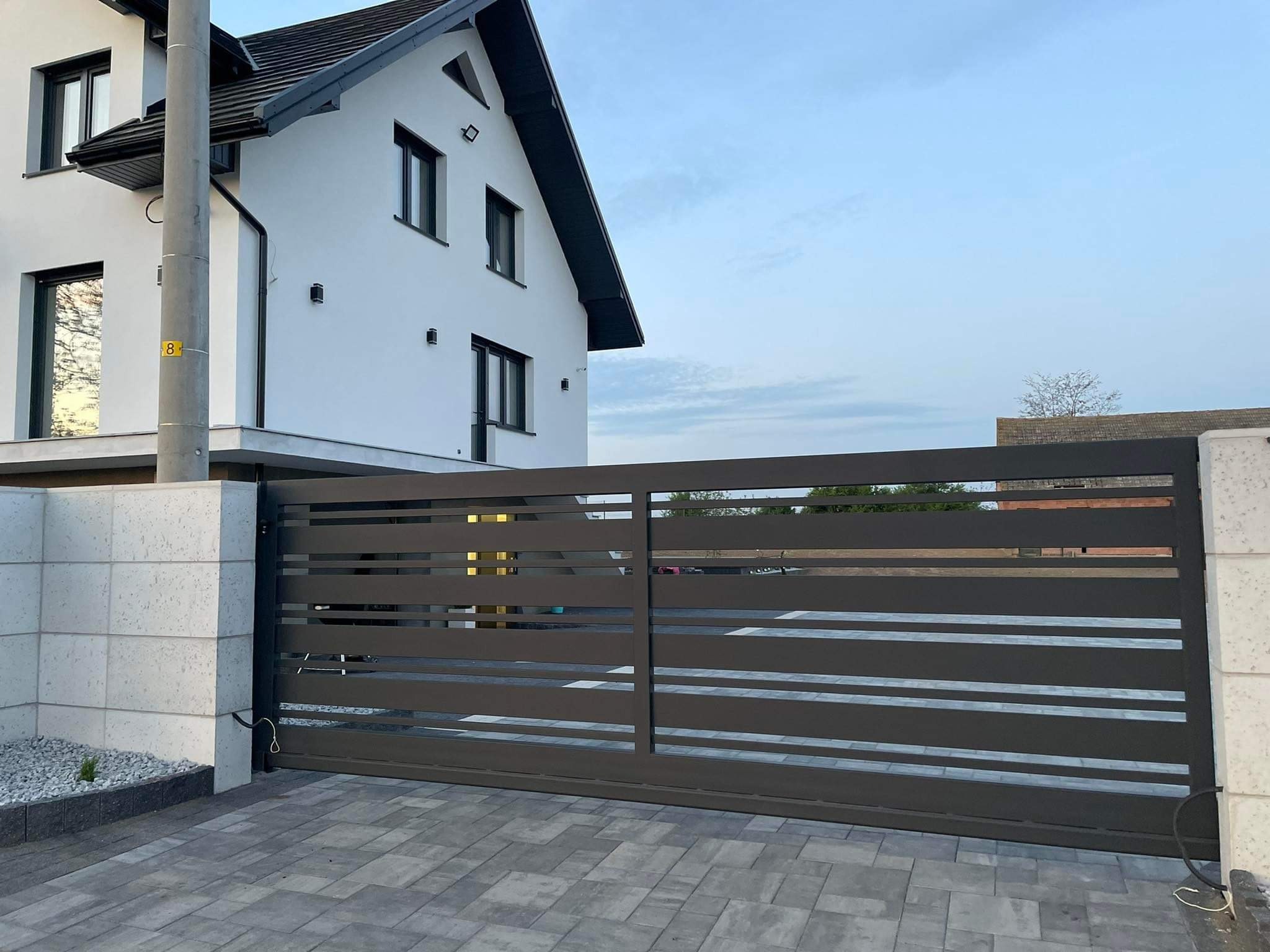 Nowoczesna, pozioma brama przesuwna w kolorze grafitowym, z widocznym kablem zasilającym, na tle białego domu z czarnym dachem i kostki brukowej.