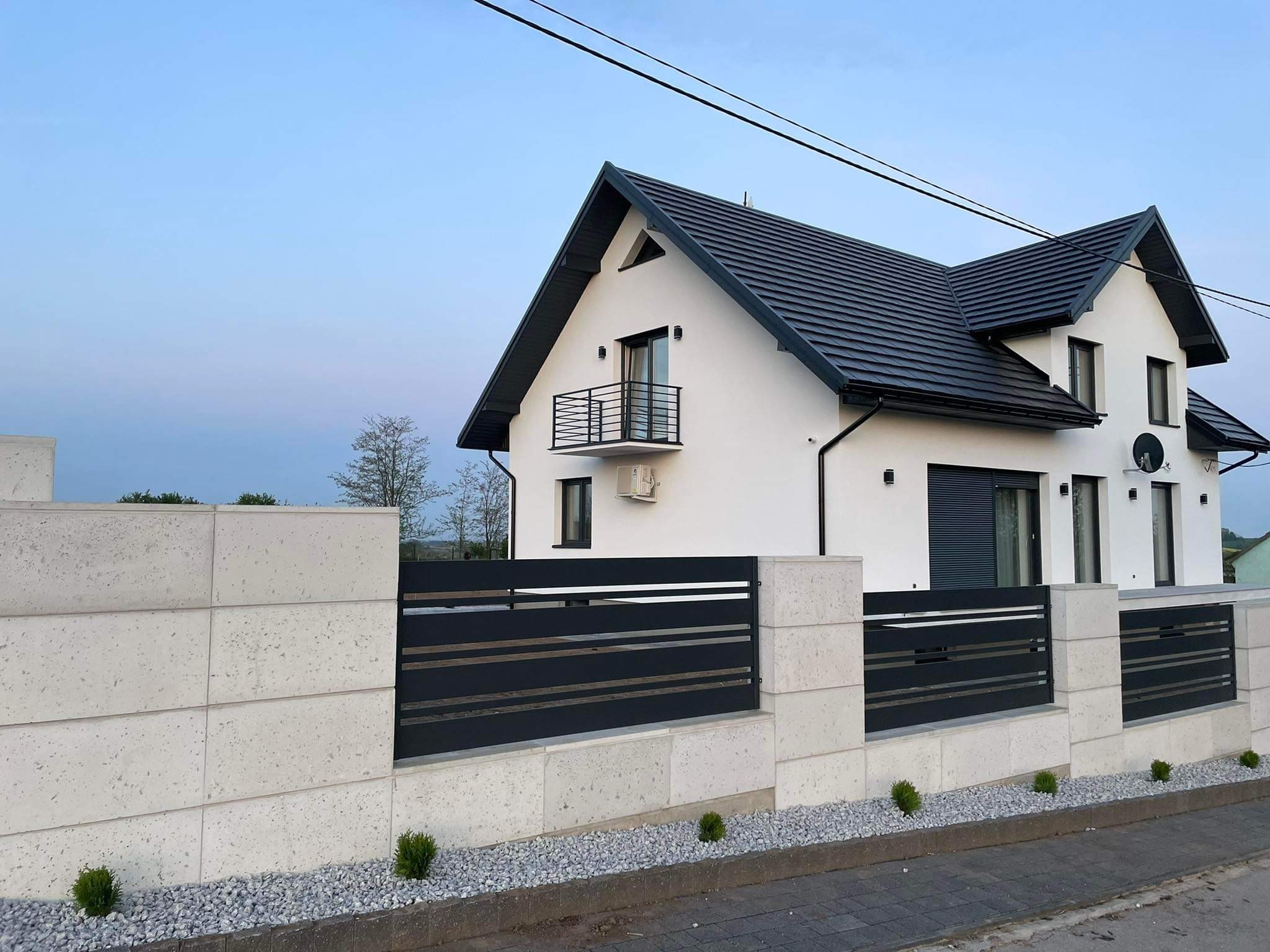 Nowoczesne ogrodzenie z poziomych, czarnych paneli metalowych osadzonych między jasnymi, kamiennymi słupkami, z eleganckim domem w tle.